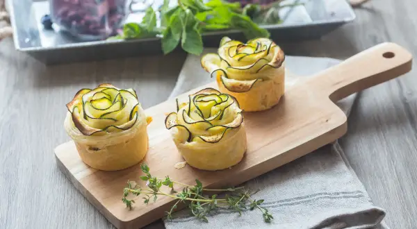 Trois petites bouchées en forme de rose, ornées de fines lamelles de fleur de courgette sur une planche en bois.
