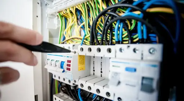 Ręka majstruje przy bezpiecznikach, gdzie kolorowe przewody symbolizują przepływ prądu elektrycznego.