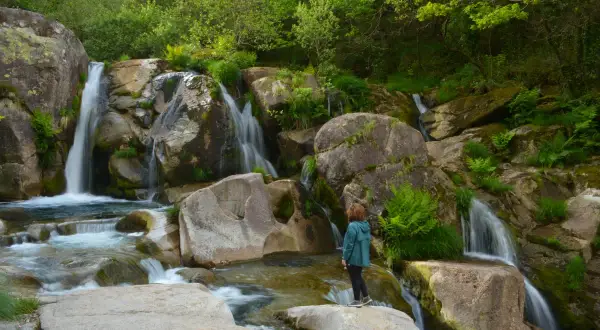 Una persona observa la hermosa fervenza de Noveira, con cascadas cayendo entre rocas cubiertas de musgo y vegetación exuberante.