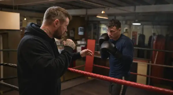 A boxer trains with his coach in a boxing ring. The coach, with tattoos on his arms and neck, demonstrates a move. The boxer, wearing gloves, listens intently. This is a scene of dedication and the pursuit of the perfect box weight.
