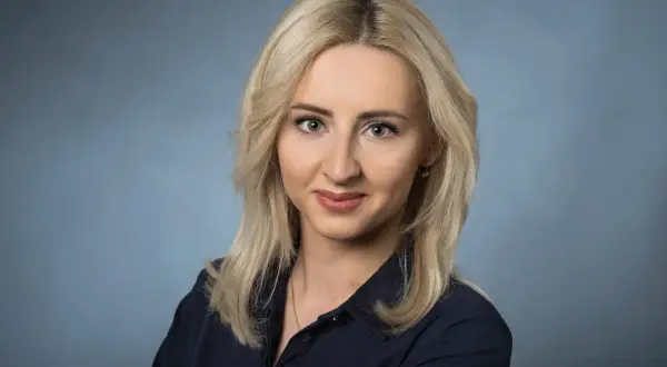 Portret uśmiechniętej blondynki w granatowej koszuli na niebieskim tle.
