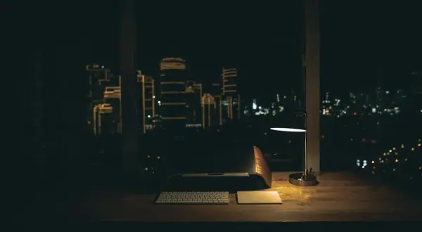 Biurko z laptopem, klawiaturą i lampką nocną. Za oknem nocne miasto, idealne do pracy w godzinach nocnych.