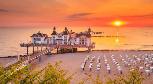 Zachód słońca nad morzem, molo z białymi budynkami, plaża z rzędami koszy plażowych. Idealne miejsce na wakacje, łatwy dojazd z Polski do Rugii.