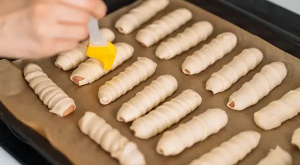 Parówki zawinięte w ciasto francuskie na blasze, przygotowywane do pieczenia.