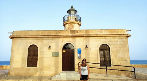 Mujer sonriente posa frente al Faro de Roquetas de Mar, con el mar azul de fondo.