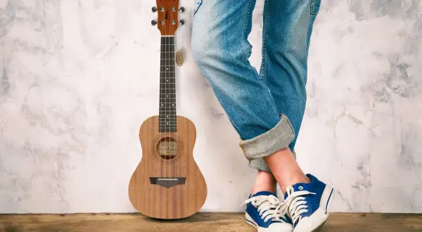 Drewniane ukulele stoi obok osoby w jeansach i niebieskich trampkach. To małe, czterostrunowe ukulele.