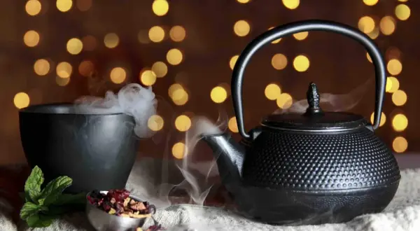 Foto still life: una teiera di ghisa nera emana vapore, accanto a una ciotola fumante e a ingredienti per il tè su uno sfondo di luci soffuse.