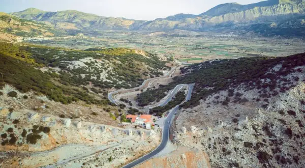 Vue aérienne d'une route sinueuse traversant un paysage montagneux verdoyant, menant à un village sur le plateau de Lassithi. Des ruines de moulins à vent parsèment la colline.