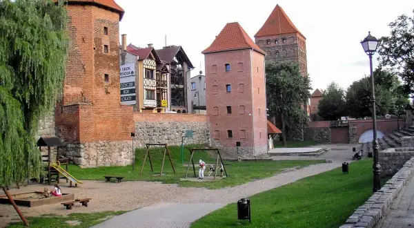 Chojnice atrakcje dla dzieci: plac zabaw przy zabytkowych wieżach. Dzieci bawią się w piaskownicy i na huśtawkach, a w tle widać ceglane budowle.
