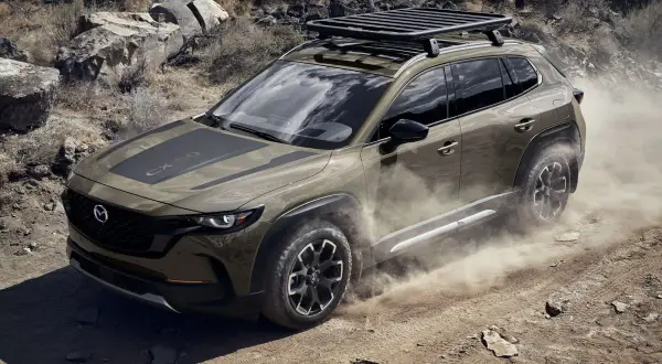 Brązowa Mazda CX-50 z bagażnikiem dachowym przemierza pustynny teren, wzbijając tumany kurzu.