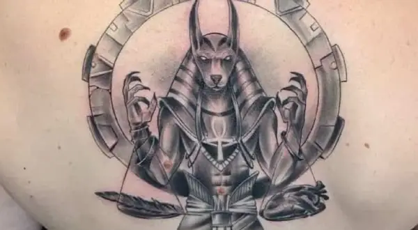 Tatuaje de Anubis con balanza, pluma y corazón, rodeado por un círculo mecánico.
