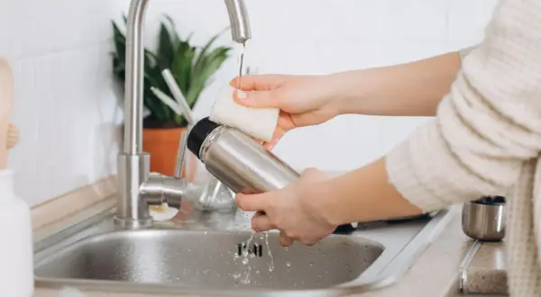 Hände reinigen eine Edelstahl-Thermoskanne unter fließendem Wasser im Spülbecken.