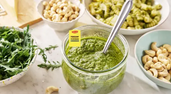 Słoik pełen aromatycznego pesto z rukoli, obok miseczki z orzechami nerkowca i świeżą rukolą. Idealne do makaronu!