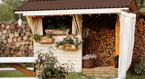 Charmante Holzunterstand Ideen: Ein gemütlicher Schuppen mit Blumenkästen, Lichterketten und einem Stapel Brennholz.