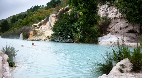 Bagno Vignoni: cascate naturali con acqua azzurra e vegetazione lussureggiante. Due persone nuotano nelle terme.