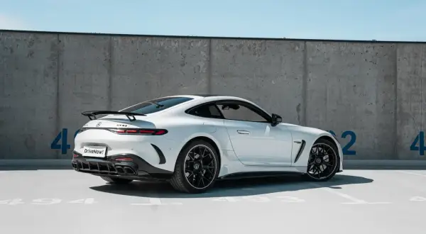 Biały Mercedes GT 63 AMG z czarnym spoilerem i felgami, zaparkowany pod betonową ścianą z niebieskimi cyframi.