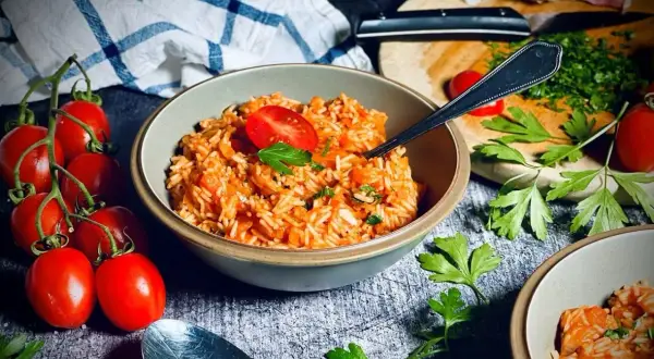 Schüssel mit aromatischem griechischem Reis, garniert mit Tomaten und Petersilie. Frische Tomaten und Knoblauch im Hintergrund.