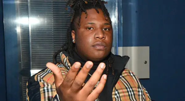 A young man with dreadlocks, wearing a plaid jacket, gestures forward. He's the artist behind many popular Tay Keith songs.