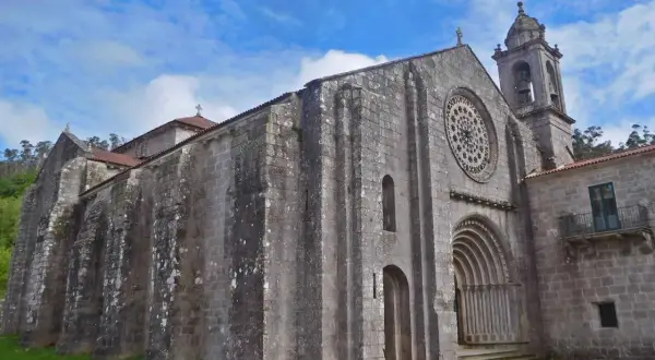 El monasterio de Armenteira, con su imponente fachada de piedra y su rosetón, se alza bajo un cielo azul con nubes.