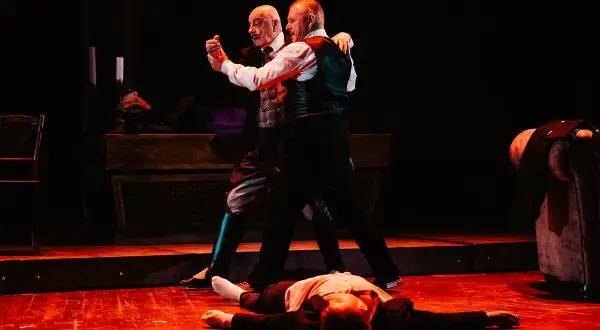 Dwóch mężczyzn tańczy tango, trzeci leży na podłodze.