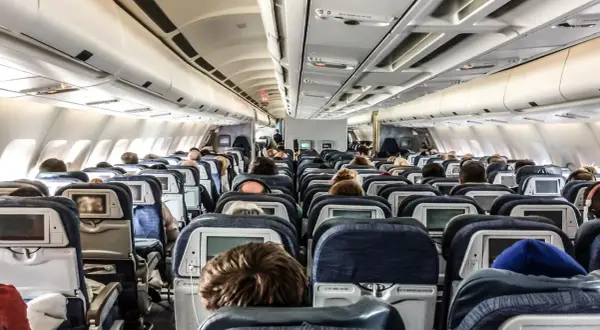 Blick in den Gang eines Flugzeugs. Viele Passagiere sitzen auf ihren Plätzen im Flugzeug innen, einige schauen auf die Bildschirme vor sich.