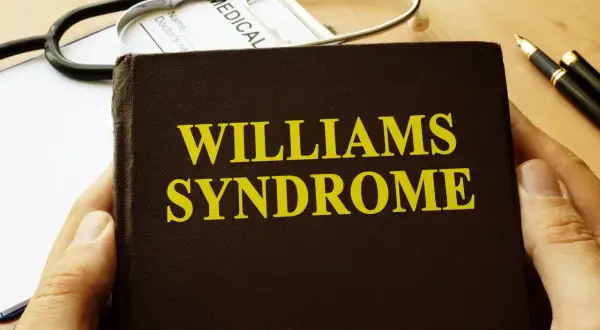 Dłoń trzyma książkę z napisem "Williams Syndrome". Obok leży stetoskop i długopis.