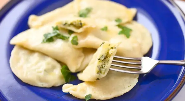 Pyszne pierogi ze szpinakiem, serem i czosnkiem, udekorowane natką pietruszki, na granatowym talerzu. Jedna porcja jest przekrojona widelcem.