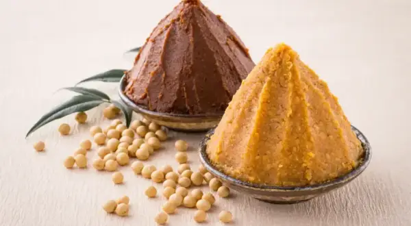 Dwa stożki pasty miso, jasnej i ciemnej, w miseczkach, obok ziaren soi i liści. Co to miso?