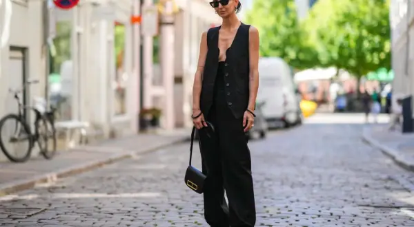 Eine Frau trägt ein festliches Oberteil zu schwarzer Hose, Sonnenbrille und einer kleinen schwarzen Tasche.