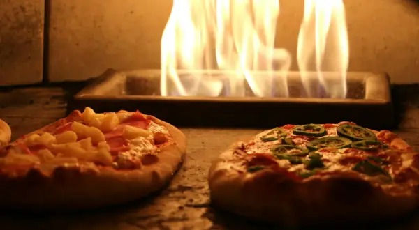 Deux pizzas maison, une à l'ananas et jambon, l'autre aux poivrons verts, cuisent dans un four avec des flammes vives. Une recette pizza maison avec pâte toute prête facile et rapide.