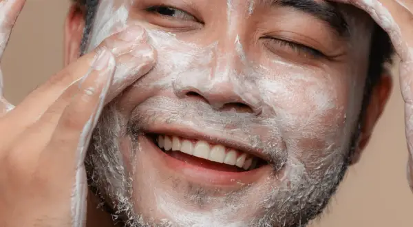 Un visage d'homme souriant, couvert de mousse nettoyante, se fait un soin du visage.