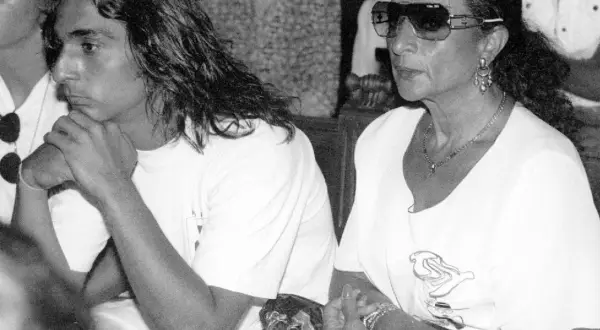 Un joven con pelo largo y una mujer con gafas de sol y joyas, sentados juntos. La imagen evoca la tristeza y el misterio en torno a como murio Lola Flores.