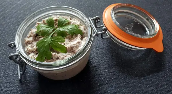 Un bocal ouvert révèle de délicieuses rillettes de sardines, garnies d'une feuille de persil frais.