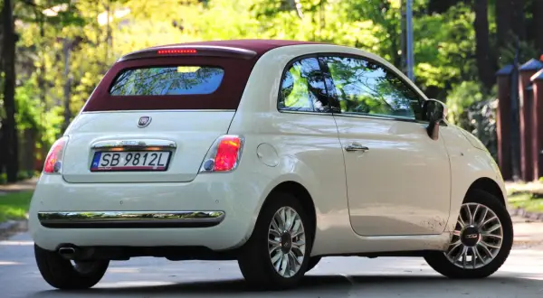 Biały fiat 500c z bordowym dachem stoi na drodze, otoczony zielenią drzew.
