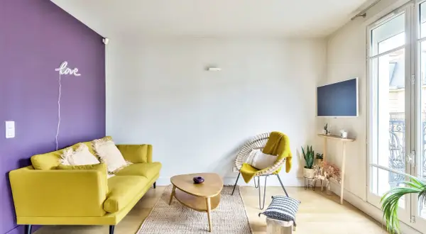 Ein gelbes Sofa und ein Korbsessel mit gelber Decke harmonieren perfekt mit der lila Wand. Welche Farbe passt zu lila? Hier sieht man, dass Gelb eine tolle Wahl ist!