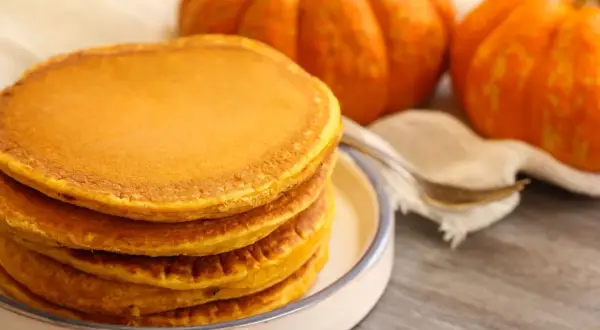 Pila de torrijas de calabaza doradas y esponjosas, con calabazas decorativas al fondo.