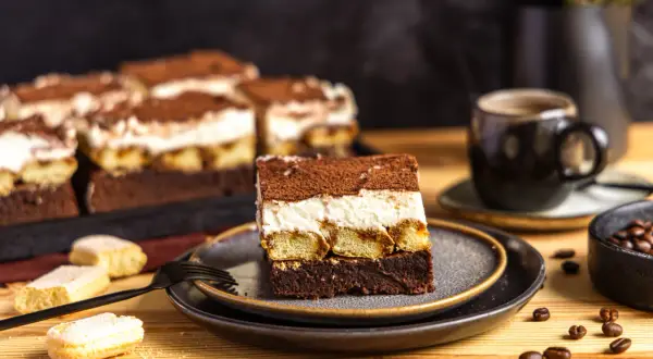 Ein Stück Tiramisu Brownie mit Löffelbiskuits, cremiger Füllung und Kakaopulver. Daneben eine Tasse Kaffee und Kaffeebohnen.