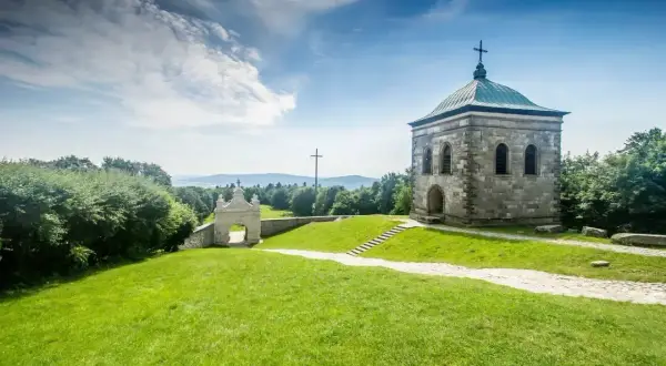 Kamienny budynek z krzyżem na dachu i krzyżem w oddali na wzgórzu. Droga prowadzi do bramy. Święty Krzyż w pełnej krasie.