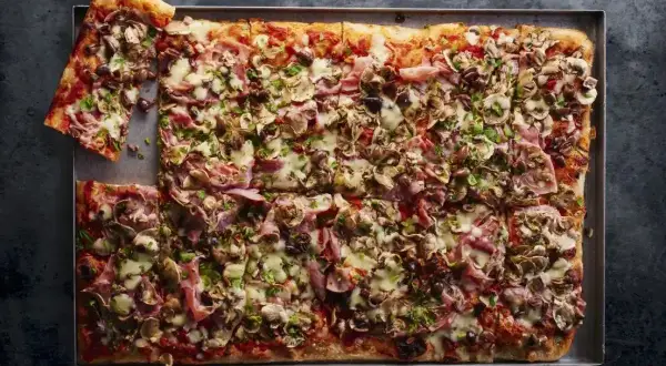 Przepis na pizzę domową na blachę: prostokątna pizza z szynką, pieczarkami i oliwkami, posypana natką pietruszki.