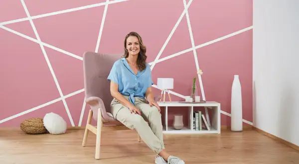 Frau sitzt vor einer Wandgestaltung mit Kreisen und Linien in Rosa und Weiß. Ein Sessel und ein Beistelltisch mit Deko stehen im Raum.