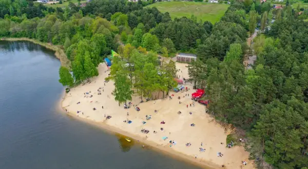 Plaża nad zalewem Cedzyna tętni życiem. Ludzie odpoczywają na piasku, ciesząc się słońcem i widokiem na jezioro.