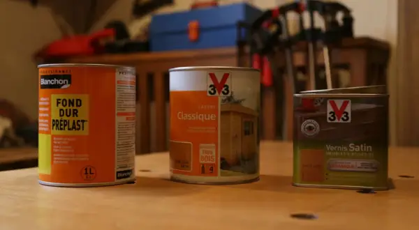 Trois boîtes de produits pour bois : un fondur avant vernis Blanchon, une lasure Classique V33 et un vernis Satin V33.