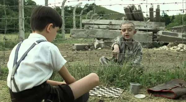Chłopiec w pasiastej koszuli wskazuje na planszę do gry. Obok siedzi inny chłopiec w białej koszuli i szelkach.