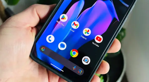 Dłoń trzyma smartfon z kolorową tapetą i ikonami aplikacji, w tym Sklep Play, Gmail, Zdjęcia i YouTube. Wygląda jak nowy android 15.