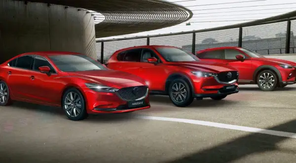 Trzy czerwone samochody marki Mazda: sedan, SUV i crossover, zaparkowane pod nowoczesną konstrukcją.