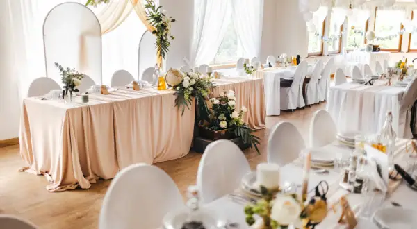 Elegancka sala weselna z białymi obrusami i kwiatami. Na stole pod dębem przywory stoi globus i walizka.