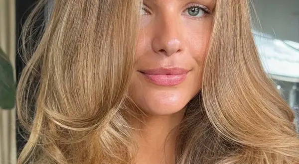Piękna kobieta z falowanymi włosami w odcieniu maślany blond. Jej oczy są zielone, a usta delikatnie umalowane.