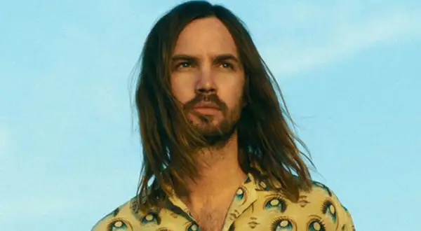 Kevin Parker w koszuli z motywem oczu, z rozwianymi włosami na tle błękitnego nieba.