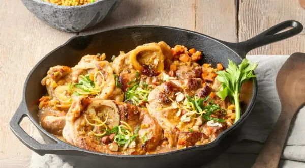 Herzhaftes Ossobuco alla Milanese in Gusseisenpfanne, garniert mit Zitronenzeste und Kräutern, daneben eine Schüssel Reis mit Erbsen.