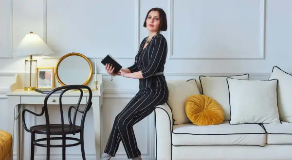 Elegancka kobieta w paski, w stylu glamour ubrania, siedzi na sofie, trzymając książkę. Obok biurko z lampą i lustrem.